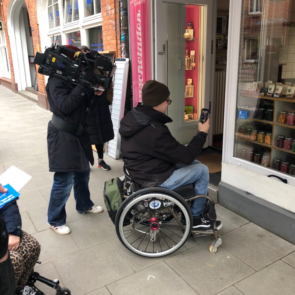 Ein Rollstuhlfahrer wird gefilmt, während er ein Foto vor dem "Bonscheladen" macht.