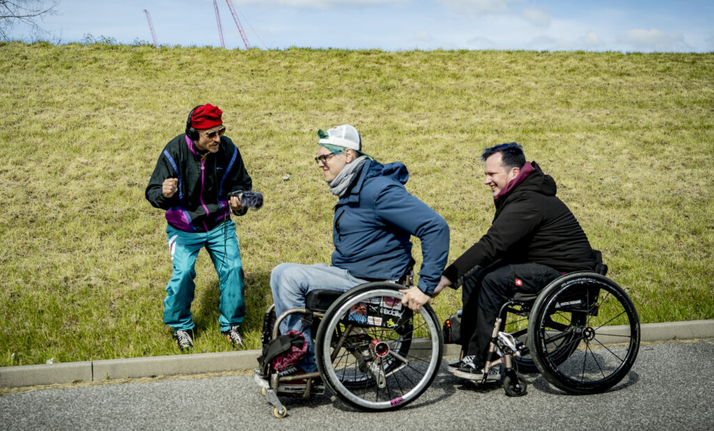 Zwei Männer fahren mit ihren Rollstühlen hintereinander und halten sich dabei fest. Ein Mann mit roter Mütze filmt das.