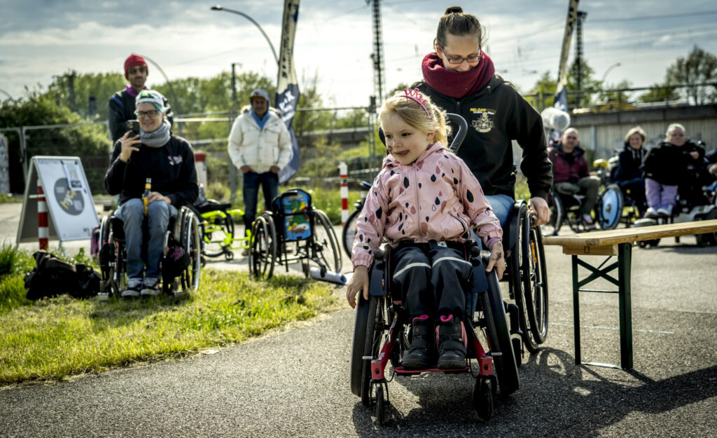 Ein Mädchen in rosaner Jacke und pinker Prinzessinnenkrone bildet mit einer erwachsenen Frau im Rollstuhl ein Tandem und fährt konzentriert die Rennstrecke.